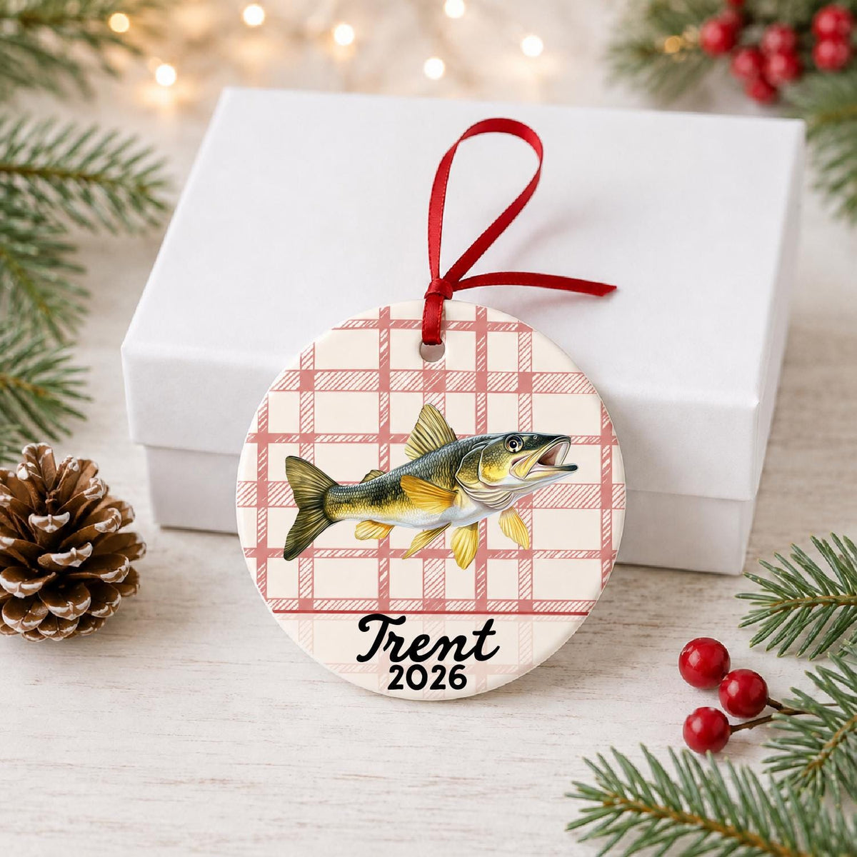 Personalized golden walleye ceramic ornament with name and year on red plaid in front of a white gift box with pinecones and red berries