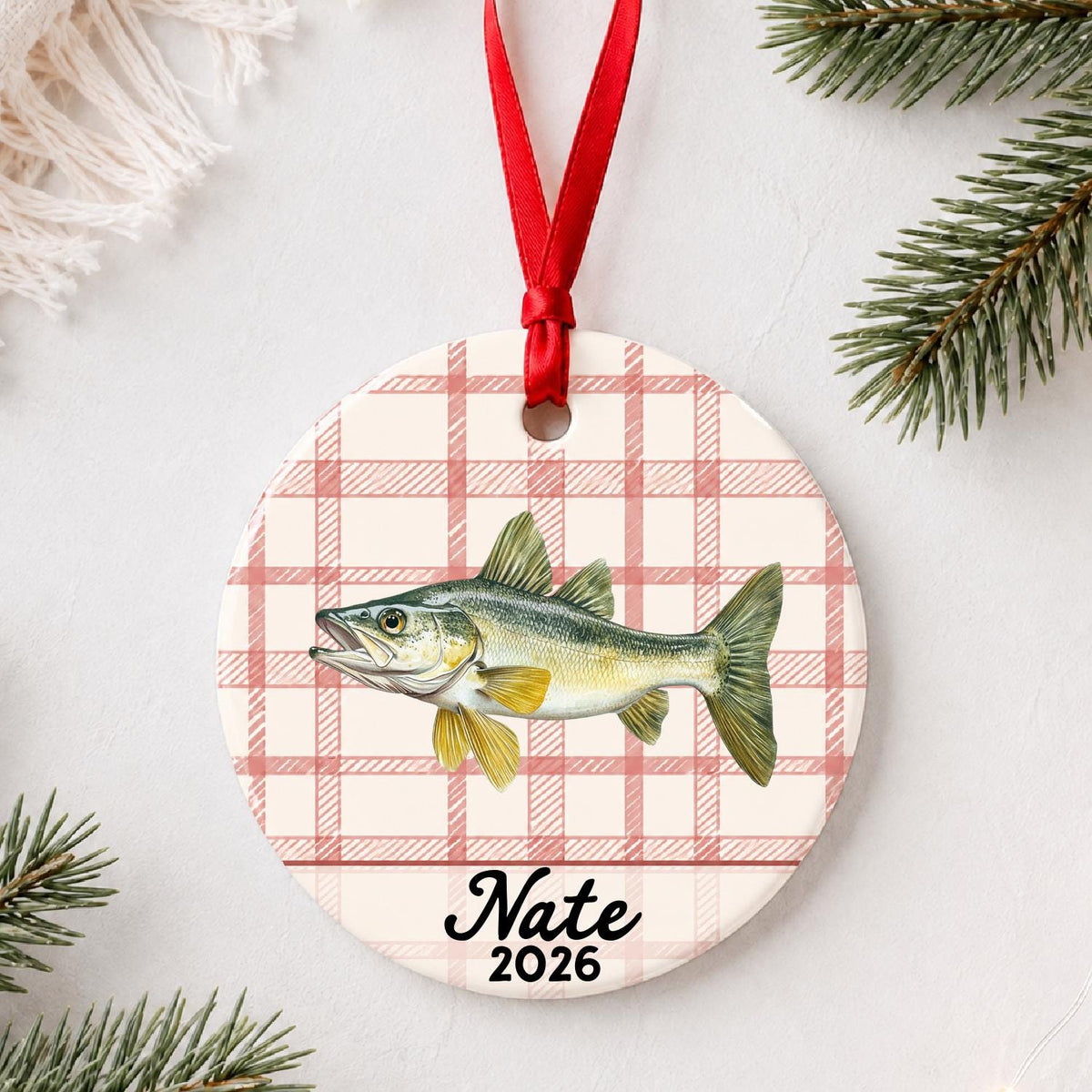 Personalized walleye ceramic ornament with name and year on red plaid styled with pine branches on a soft white background