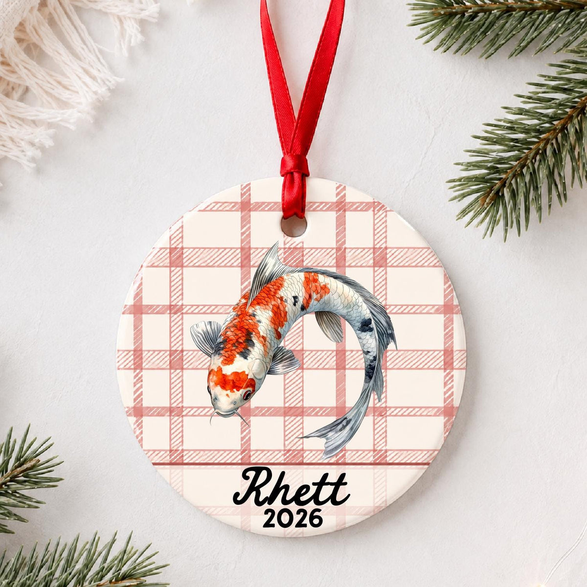Personalized koi fish ceramic ornament with name and year on red plaid styled with pine branches on a soft white background