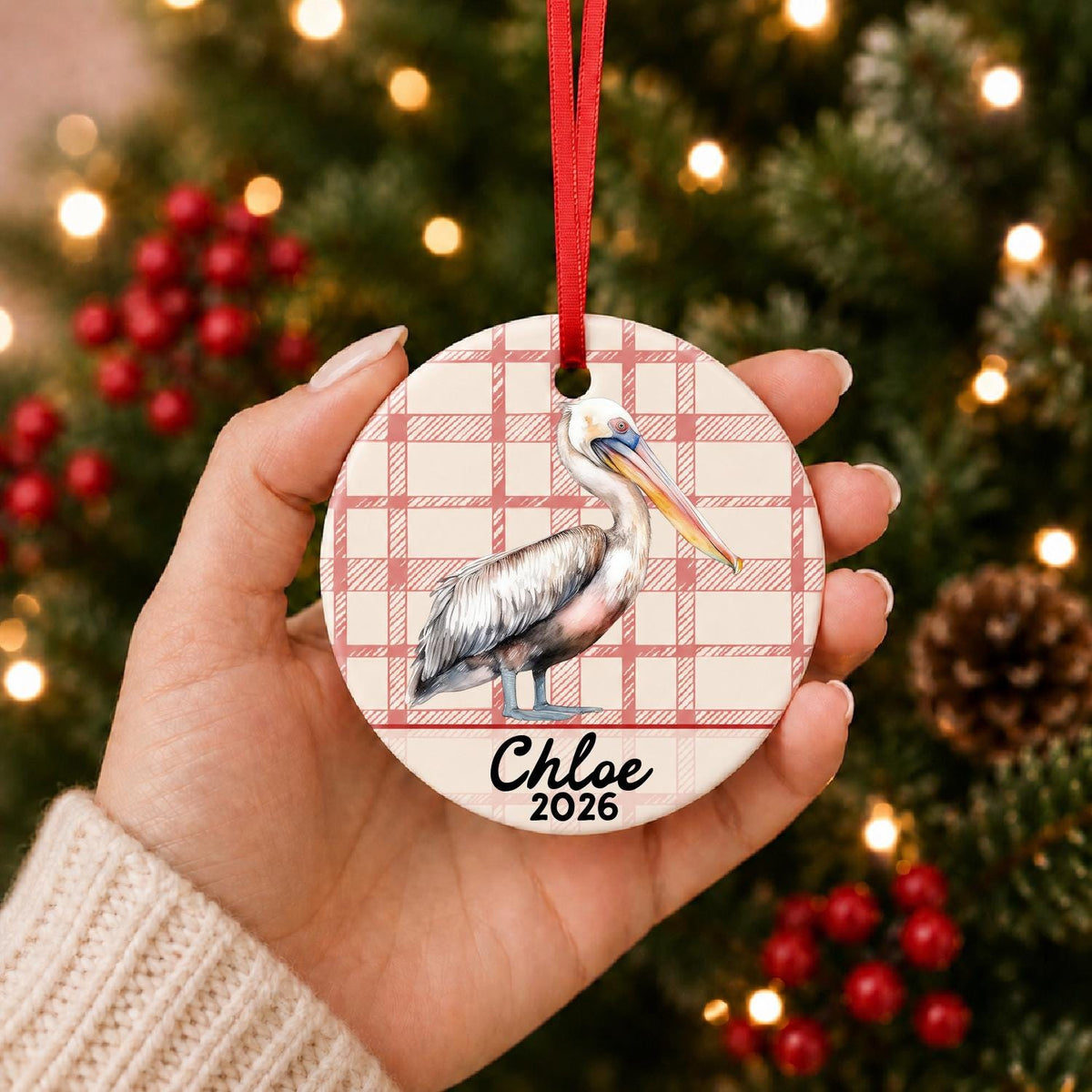 Hand holding the personalized pelican ceramic ornament with name and year on red plaid in front of a Christmas tree