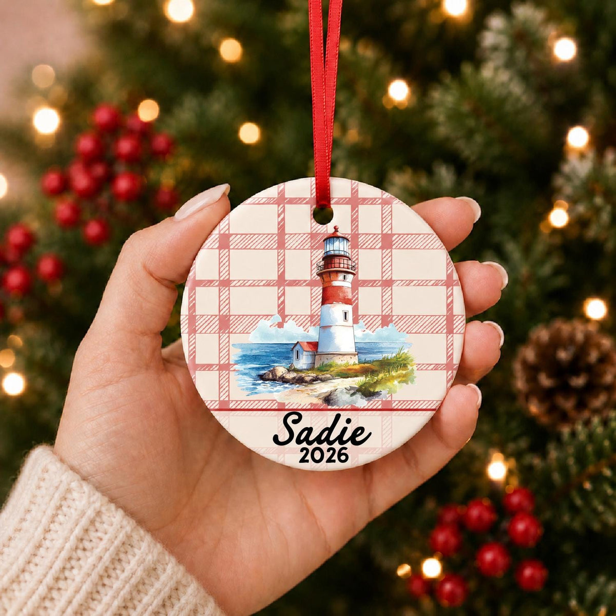Hand holding the personalized beach lighthouse ceramic ornament with name and year on red plaid in front of a Christmas tree