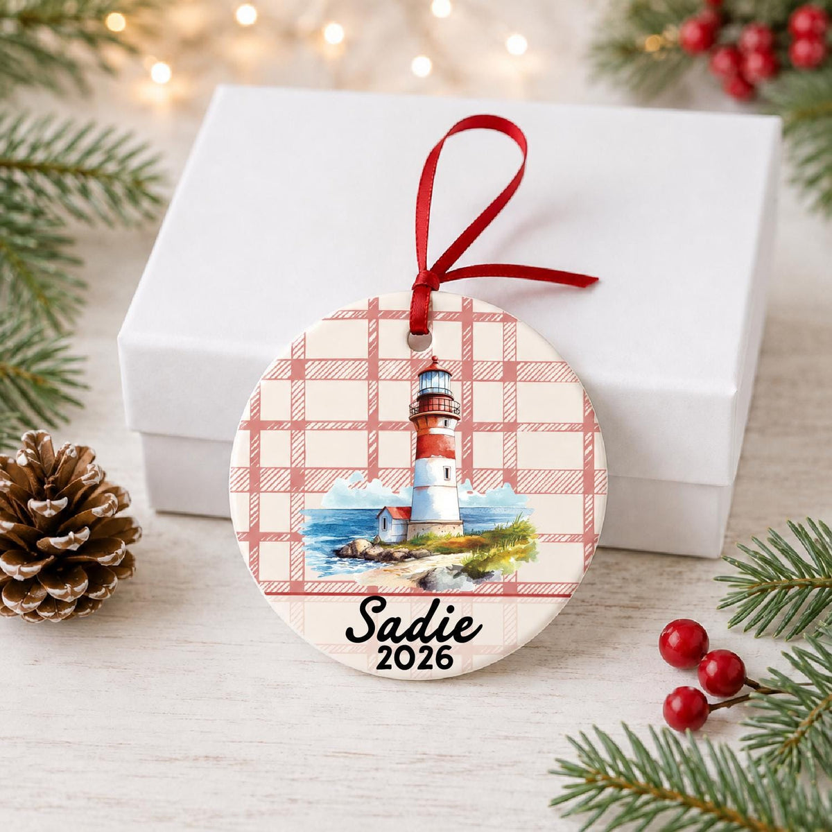 Personalized beach lighthouse ceramic ornament with name and year on red plaid in front of a white gift box with pinecones and red berries