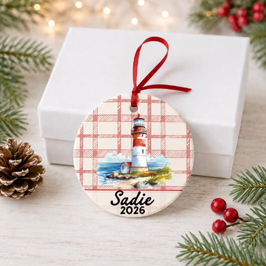 Personalized beach lighthouse ceramic ornament with name and year on red plaid in front of a white gift box with pinecones and red berries