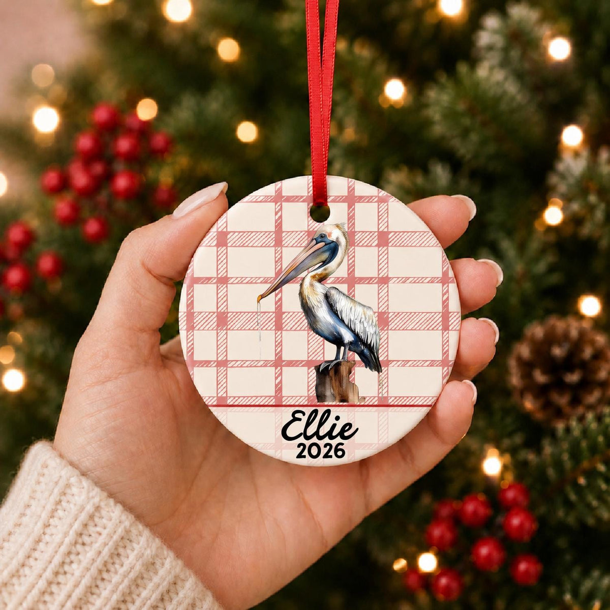 Hand holding the personalized pelican on piling ceramic ornament with name and year on red plaid in front of a Christmas tree