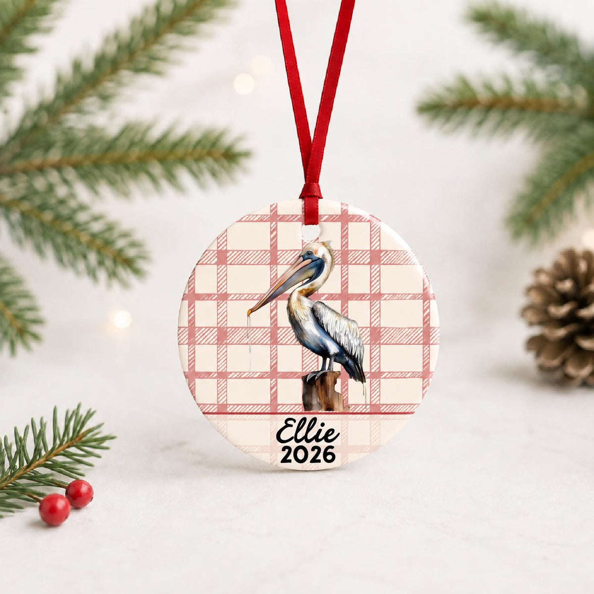 Personalized pelican on piling ceramic ornament with name and year on red plaid with red ribbon on a white surface with pine branches and pinecones
