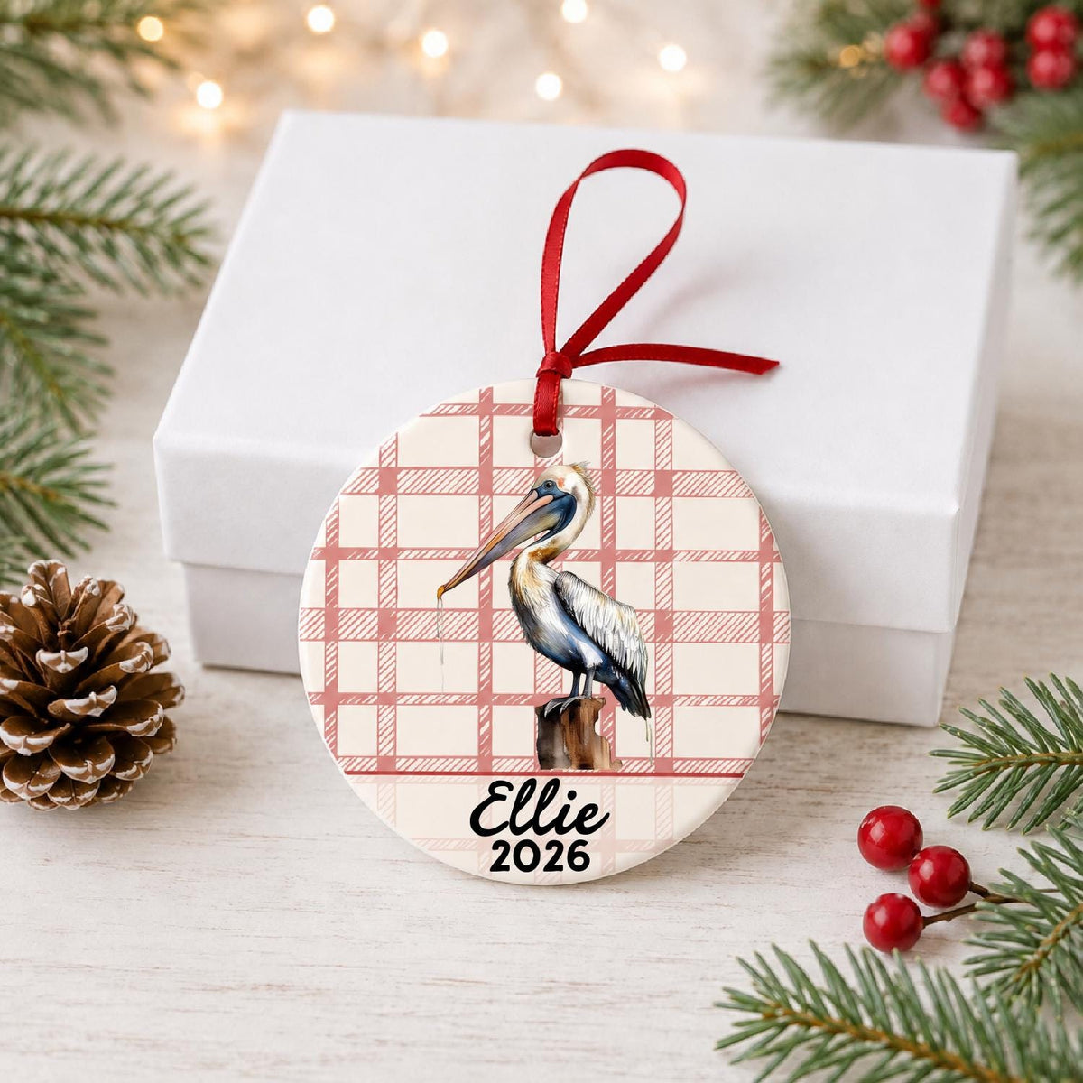 Personalized pelican on piling ceramic ornament with name and year on red plaid in front of a white gift box with pinecones and red berries