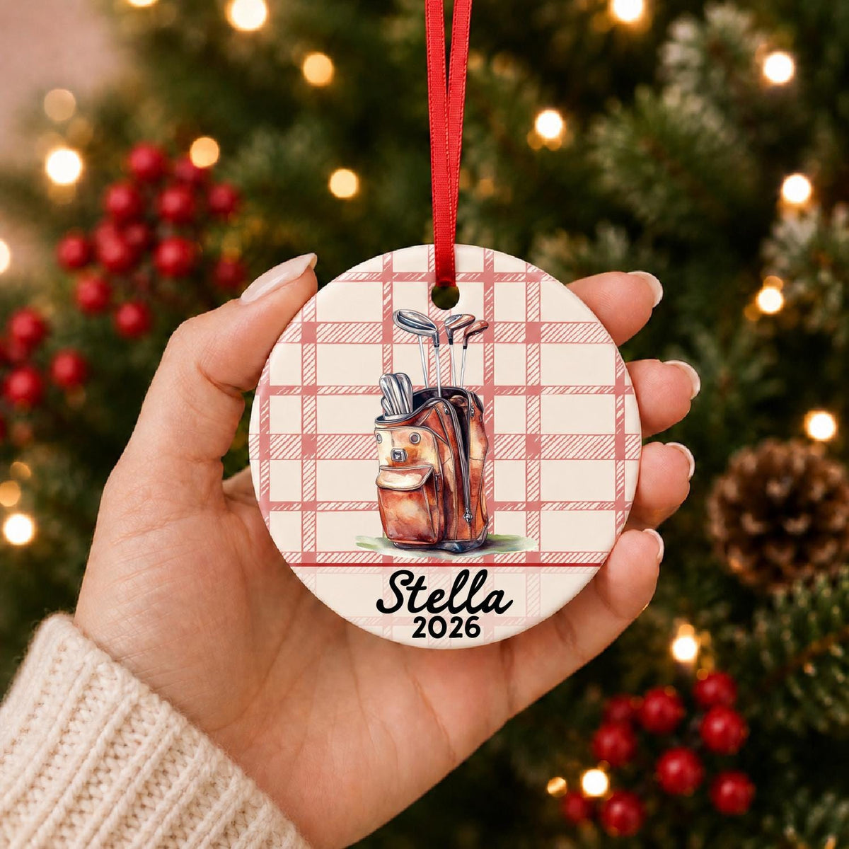 Hand holding the personalized golf bag ceramic ornament with name and year on red plaid in front of a Christmas tree