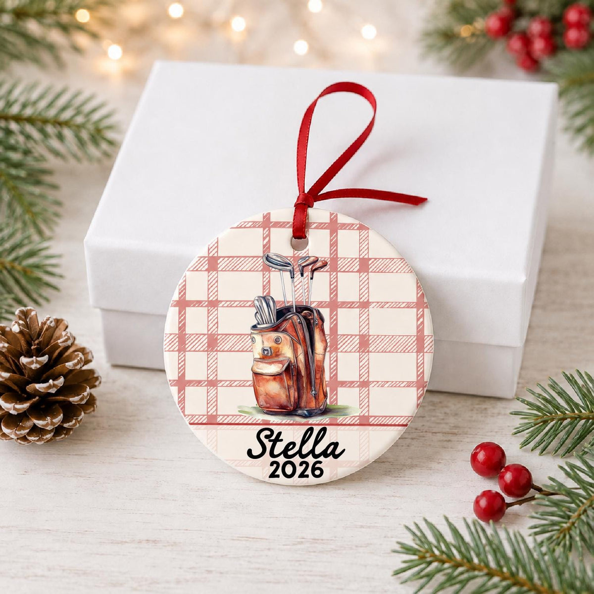 Personalized golf bag ceramic ornament with name and year on red plaid in front of a white gift box with pinecones and red berries