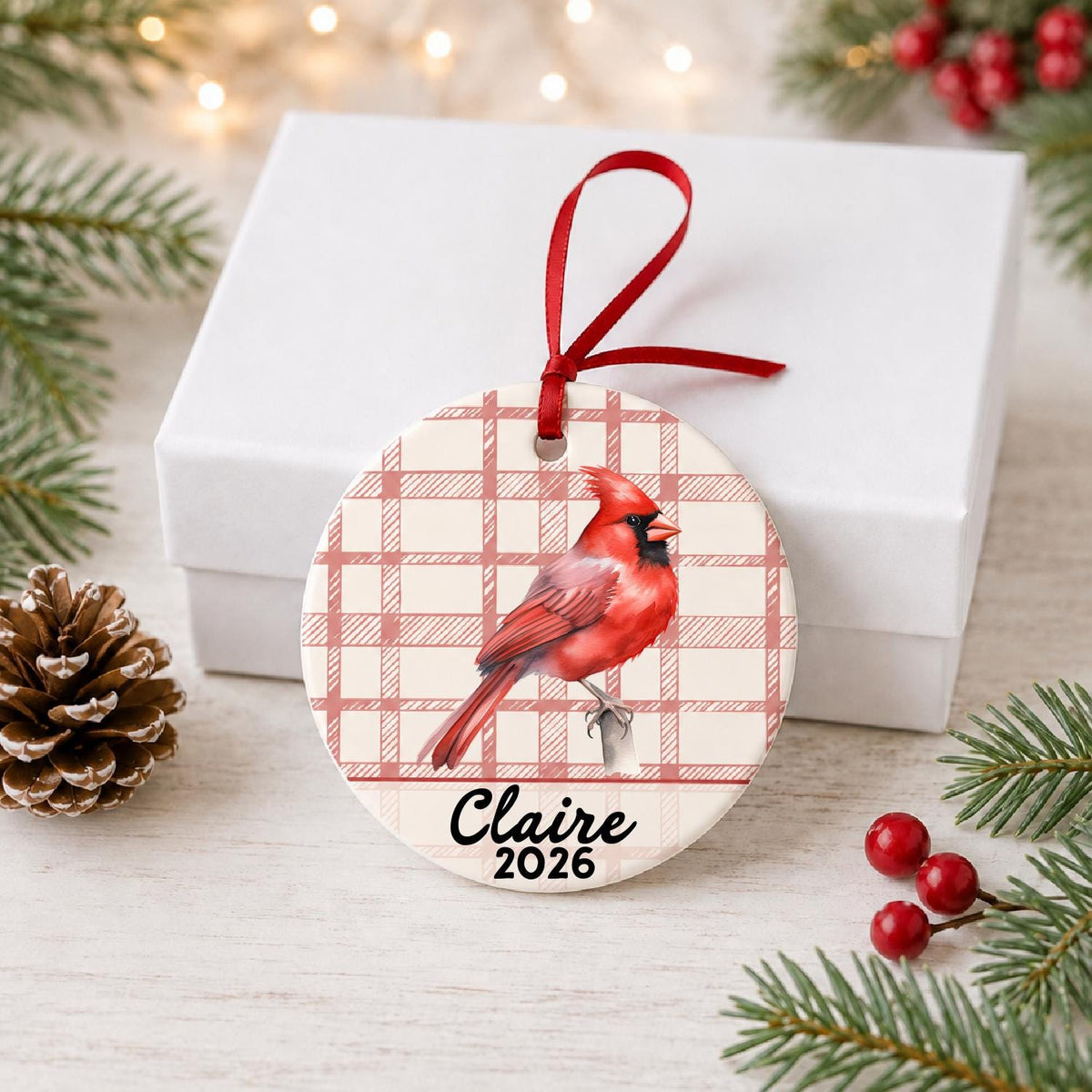 Personalized northern cardinal ceramic ornament with name and year on red plaid in front of a white gift box with pinecones and red berries