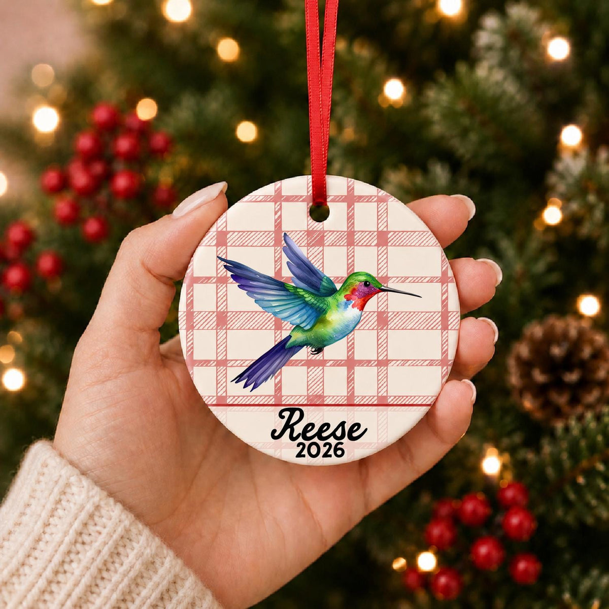 Hand holding the personalized hummingbird ceramic ornament with name and year on red plaid in front of a Christmas tree