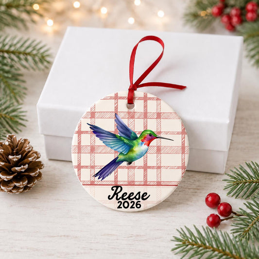 Personalized hummingbird ceramic ornament with name and year on red plaid in front of a white gift box with pinecones and red berries