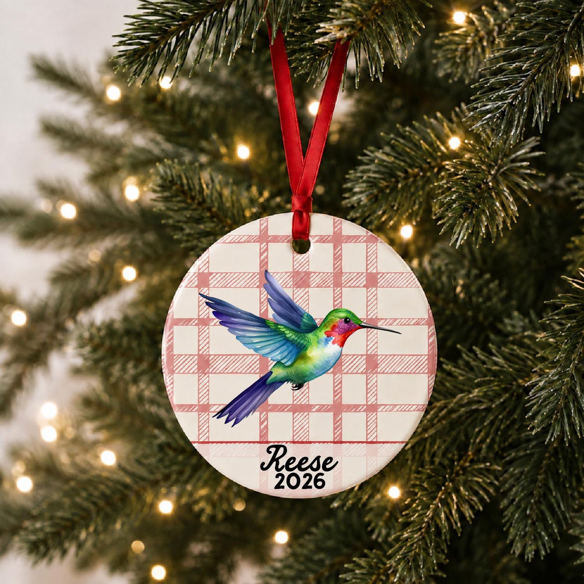 Personalized hummingbird ceramic ornament with name and year on red plaid hanging on a lit Christmas tree