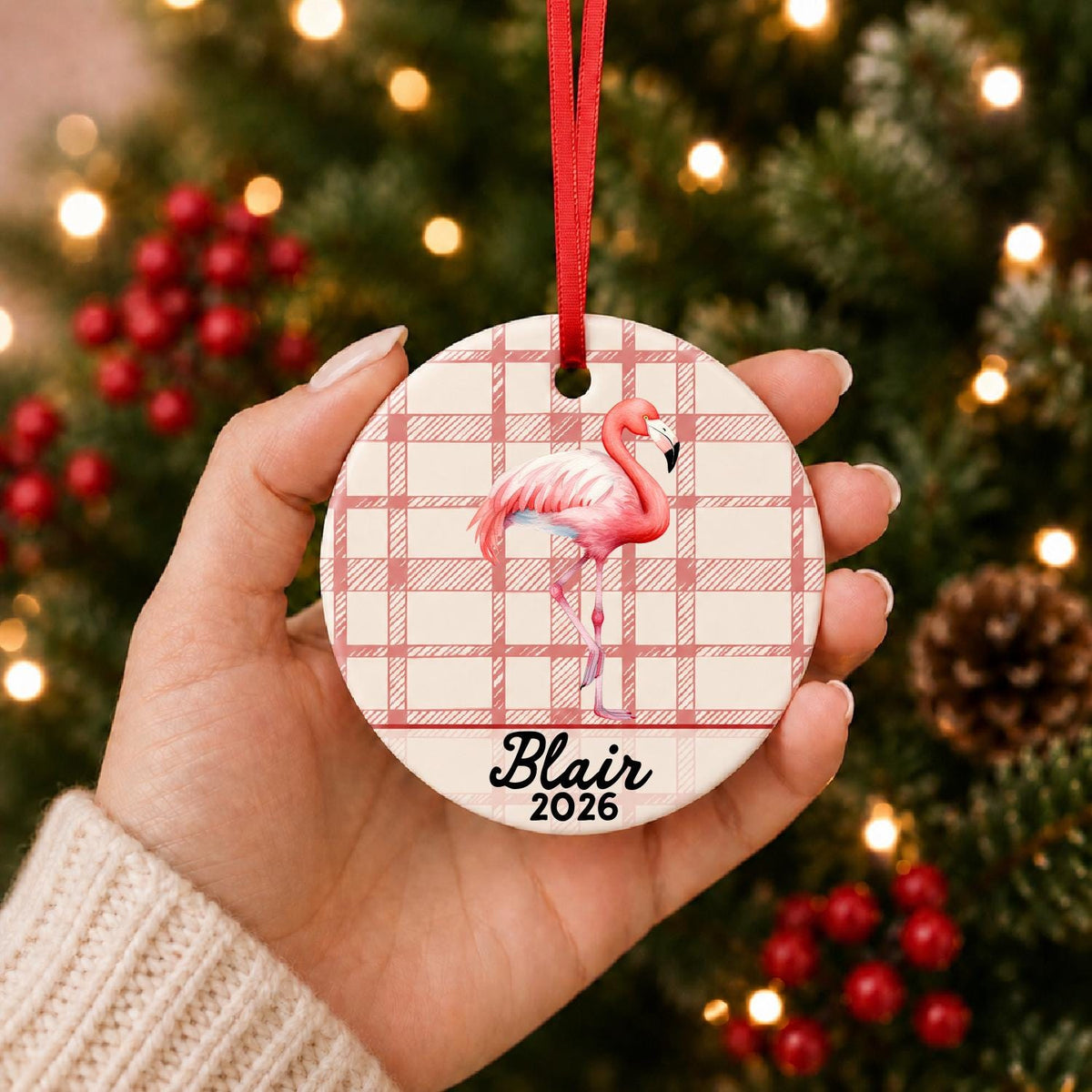 Hand holding the personalized flamingo ceramic ornament with name and year on red plaid in front of a Christmas tree
