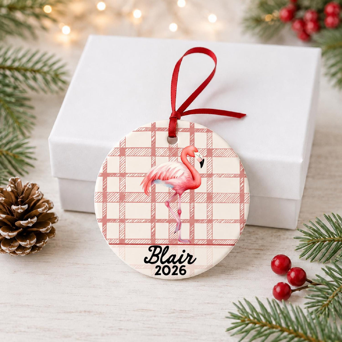 Personalized flamingo ceramic ornament with name and year on red plaid in front of a white gift box with pinecones and red berries