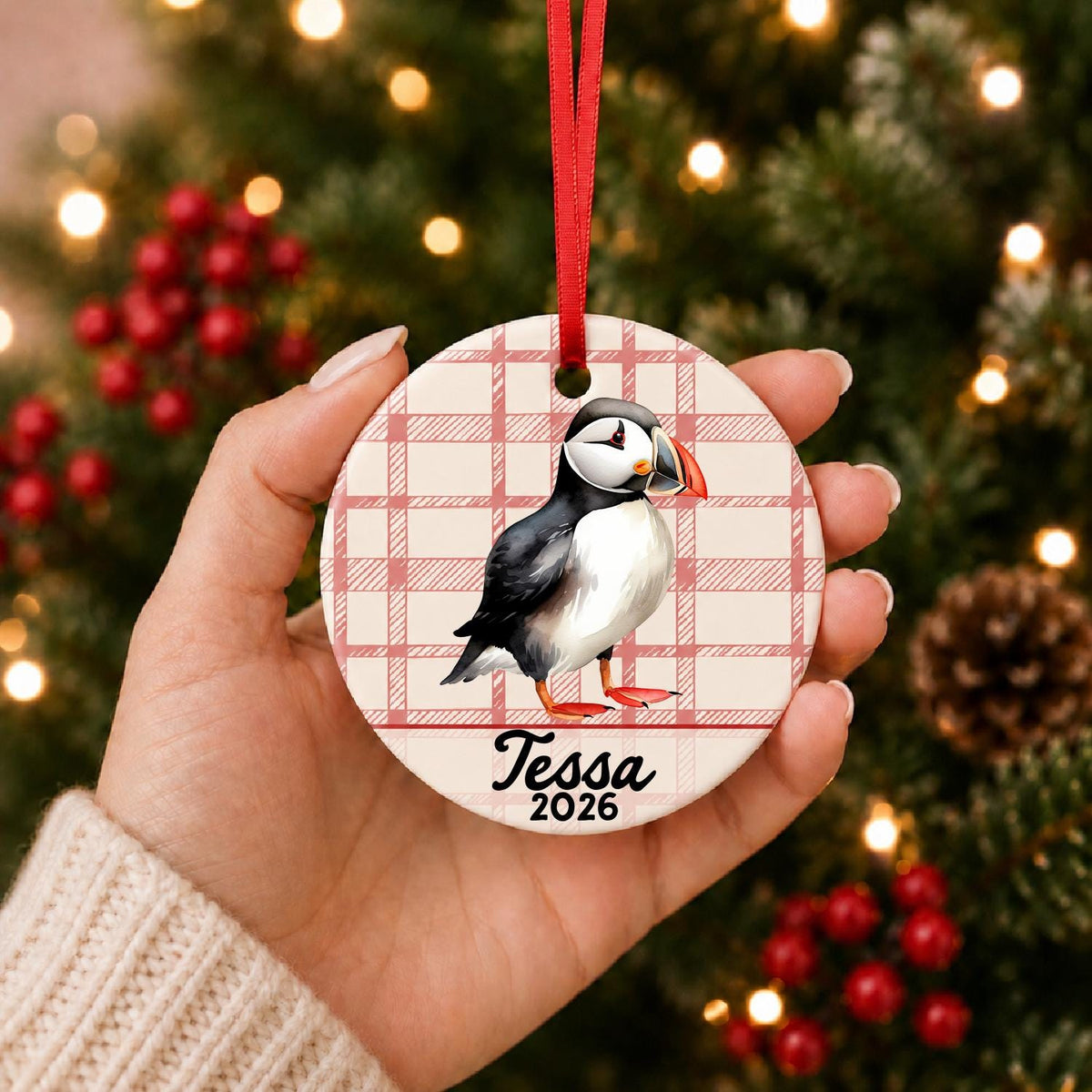 Hand holding the personalized puffin ceramic ornament with name and year on red plaid in front of a Christmas tree