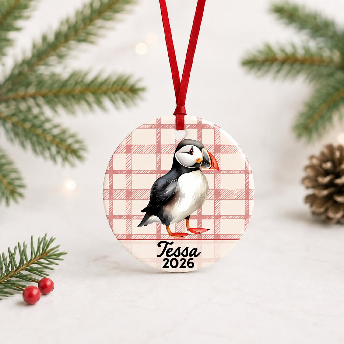 Personalized puffin ceramic ornament with name and year on red plaid with red ribbon on a white surface with pine branches and pinecones