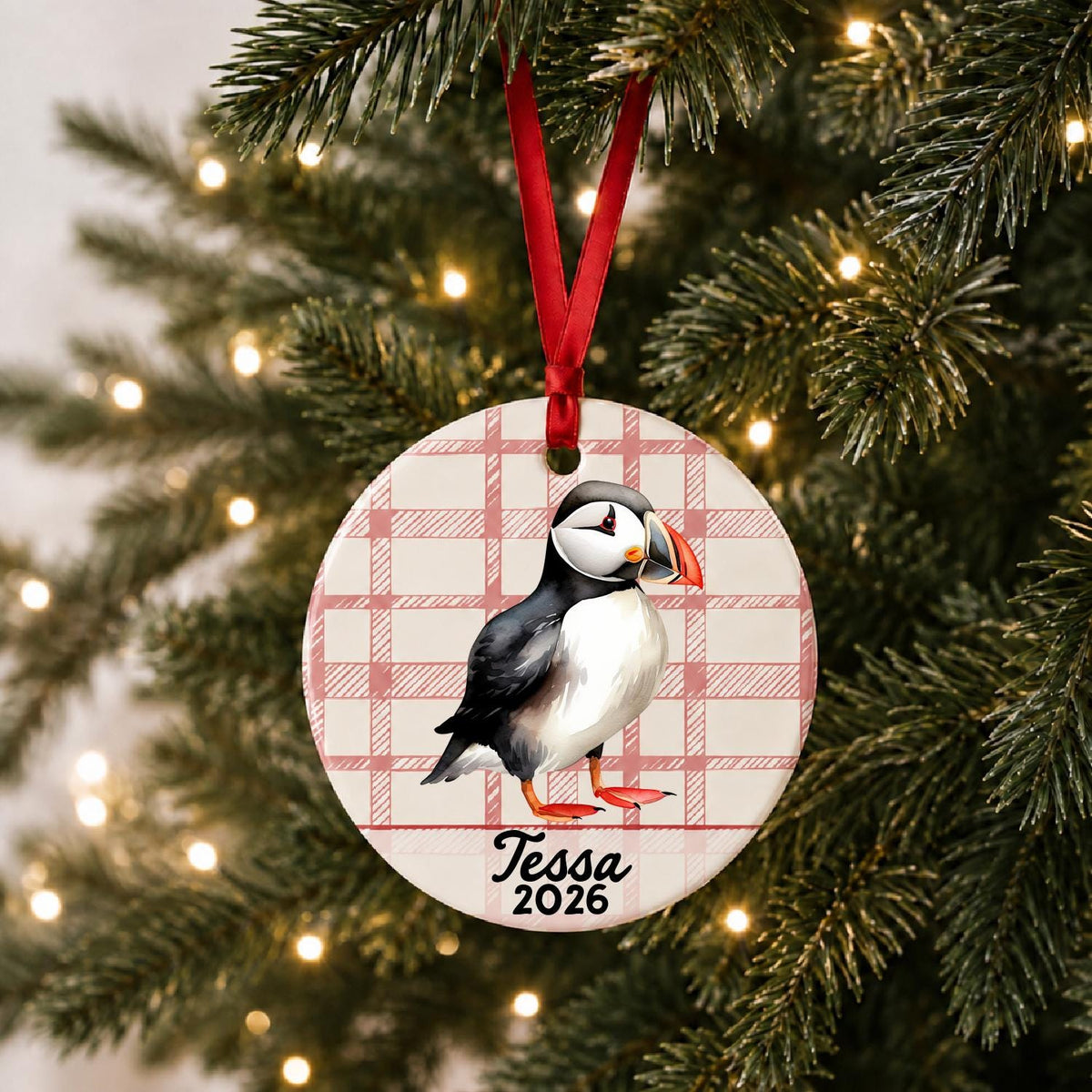 Personalized puffin ceramic ornament with name and year on red plaid hanging on a lit Christmas tree