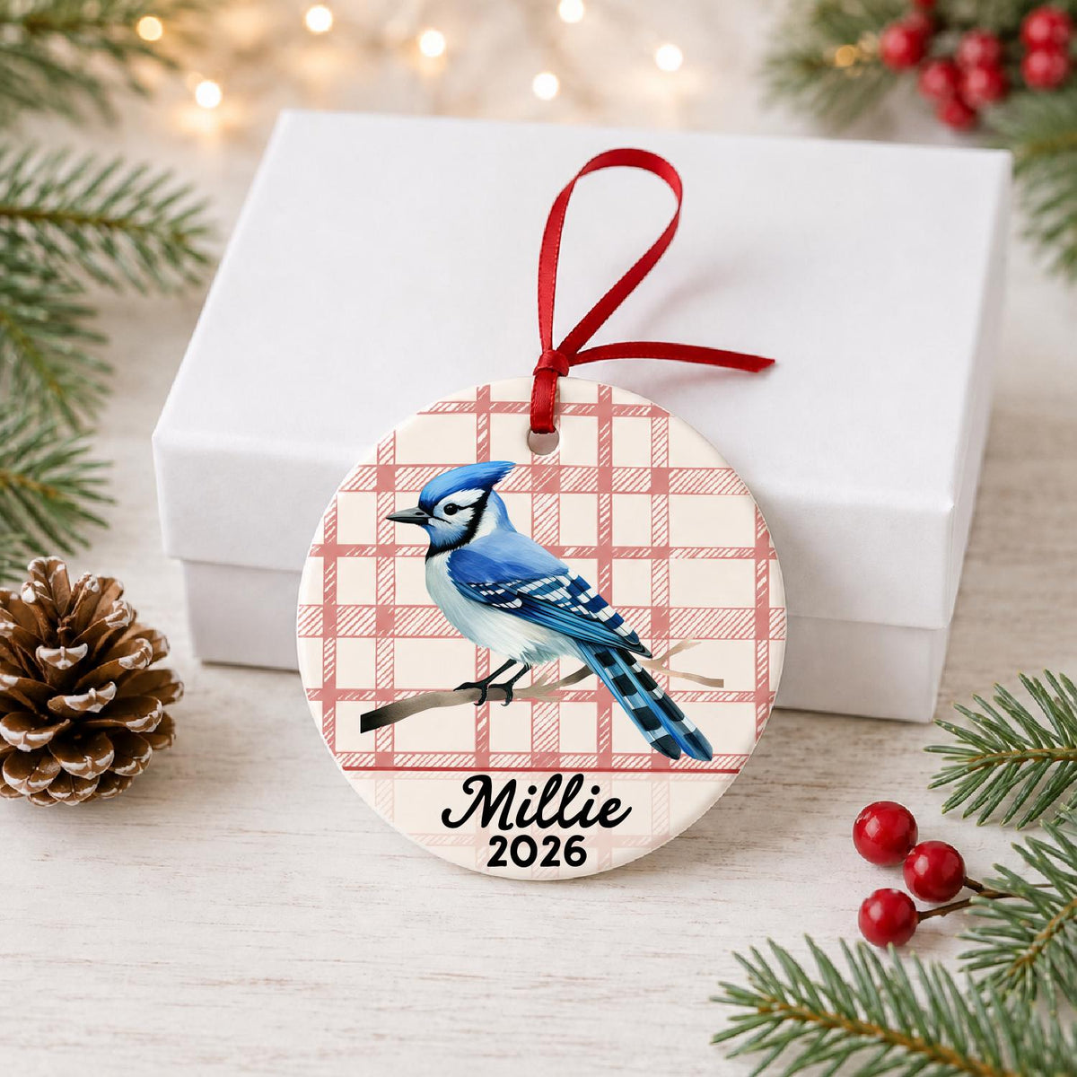 Personalized blue jay ceramic ornament with name and year on red plaid in front of a white gift box with pinecones and red berries