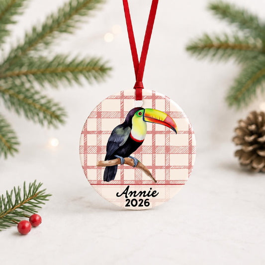 Personalized toucan ceramic ornament with name and year on red plaid with red ribbon on a white surface with pine branches and pinecones