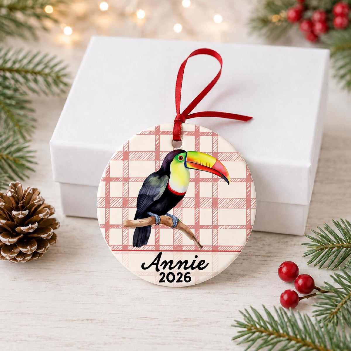 Personalized toucan ceramic ornament with name and year on red plaid in front of a white gift box with pinecones and red berries