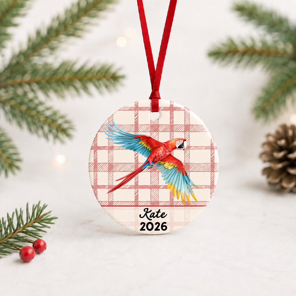 Personalized scarlet macaw ceramic ornament with name and year on red plaid with red ribbon on a white surface with pine branches and pinecones
