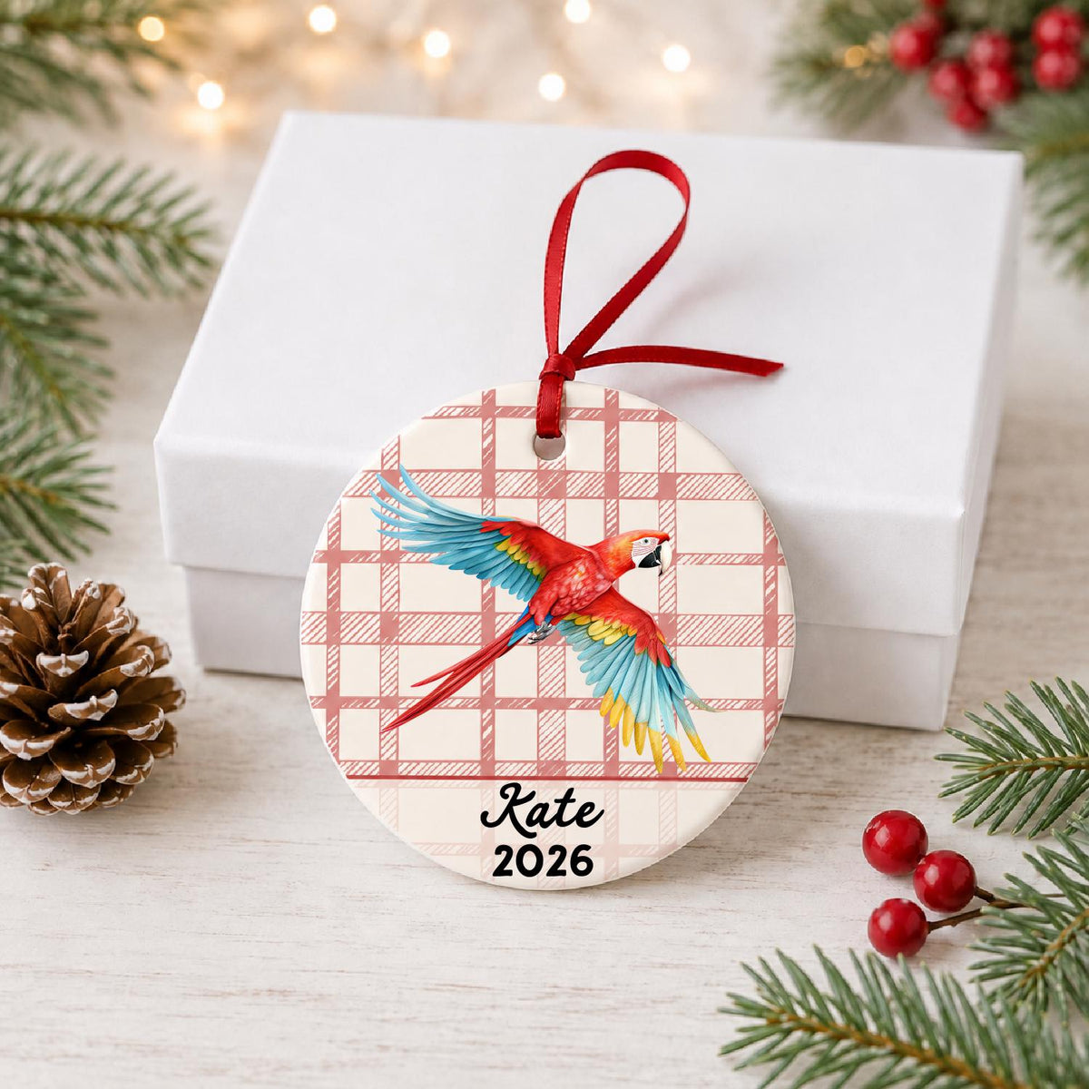 Personalized scarlet macaw ceramic ornament with name and year on red plaid in front of a white gift box with pinecones and red berries