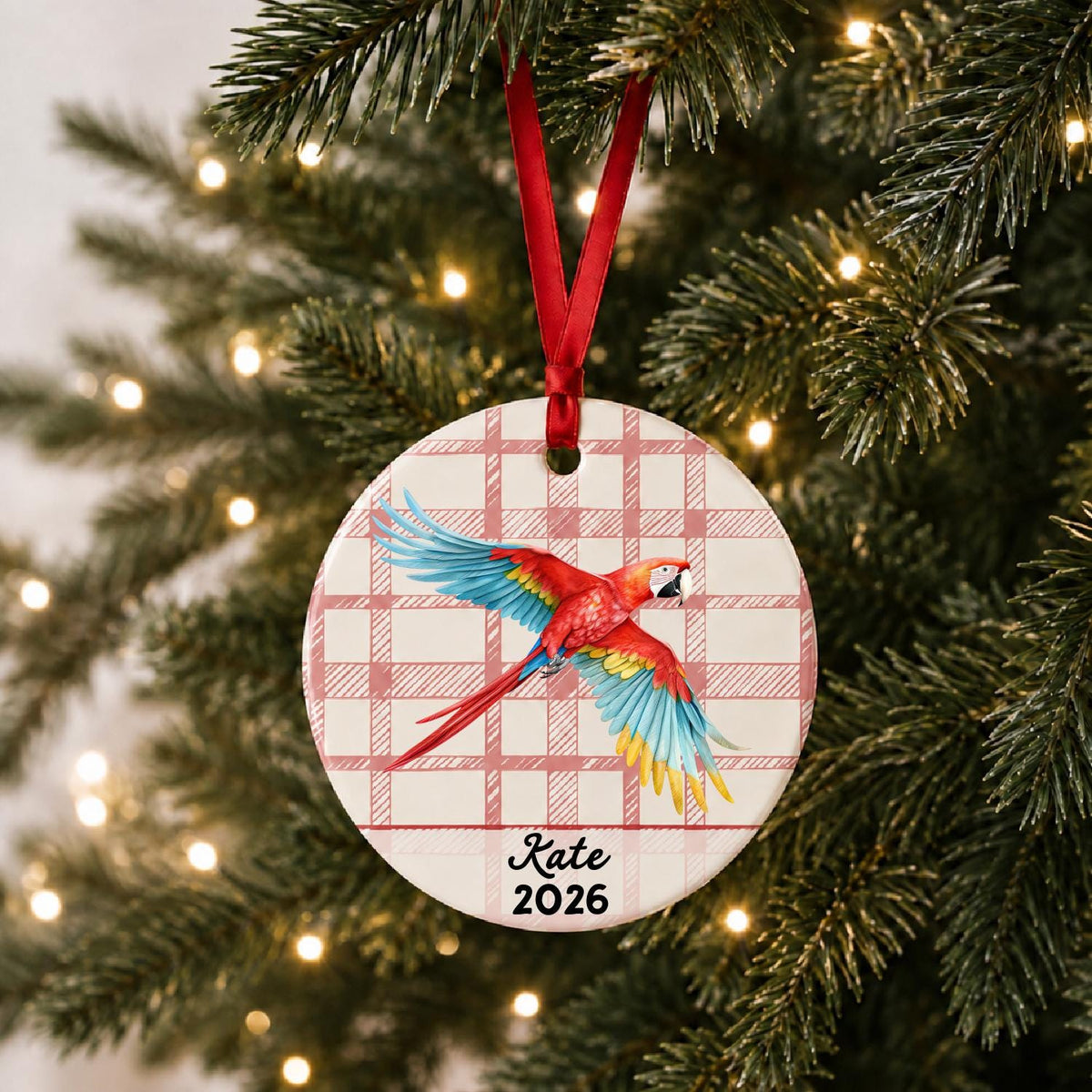 Personalized scarlet macaw ceramic ornament with name and year on red plaid hanging on a lit Christmas tree