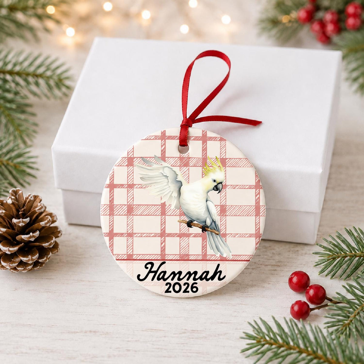 Personalized cockatoo ceramic ornament with name and year on red plaid in front of a white gift box with pinecones and red berries