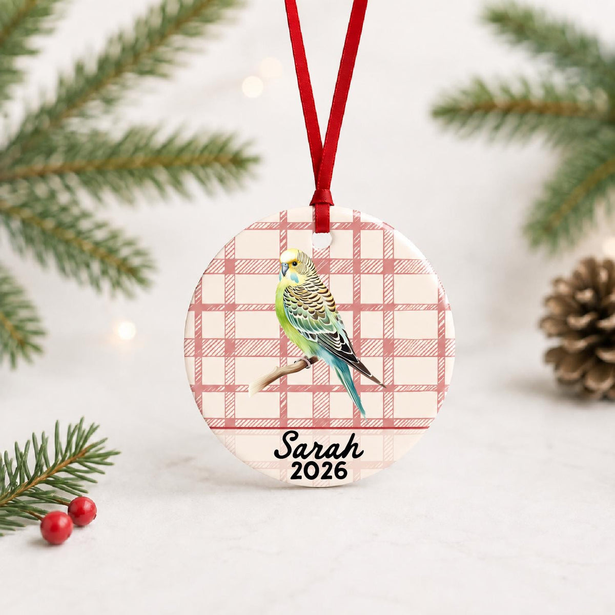 Personalized budgerigar ceramic ornament with name and year on red plaid with red ribbon on a white surface with pine branches and pinecones