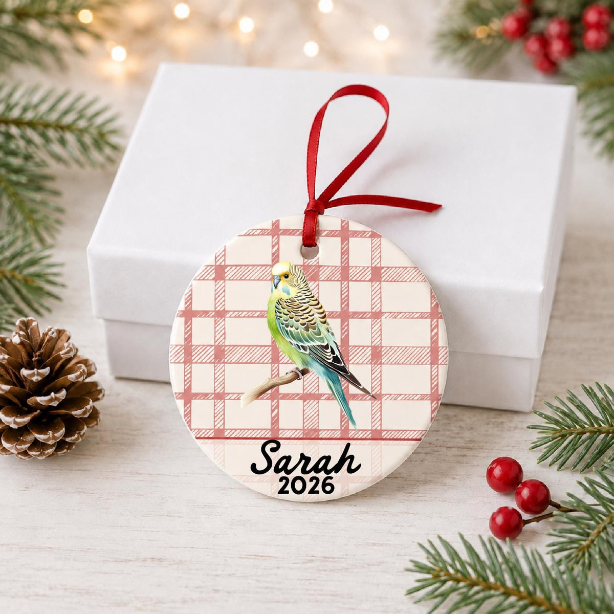 Personalized budgerigar ceramic ornament with name and year on red plaid in front of a white gift box with pinecones and red berries