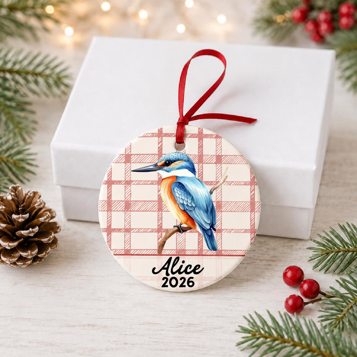 Personalized kingfisher ceramic ornament with name and year on red plaid in front of a white gift box with pinecones and red berries