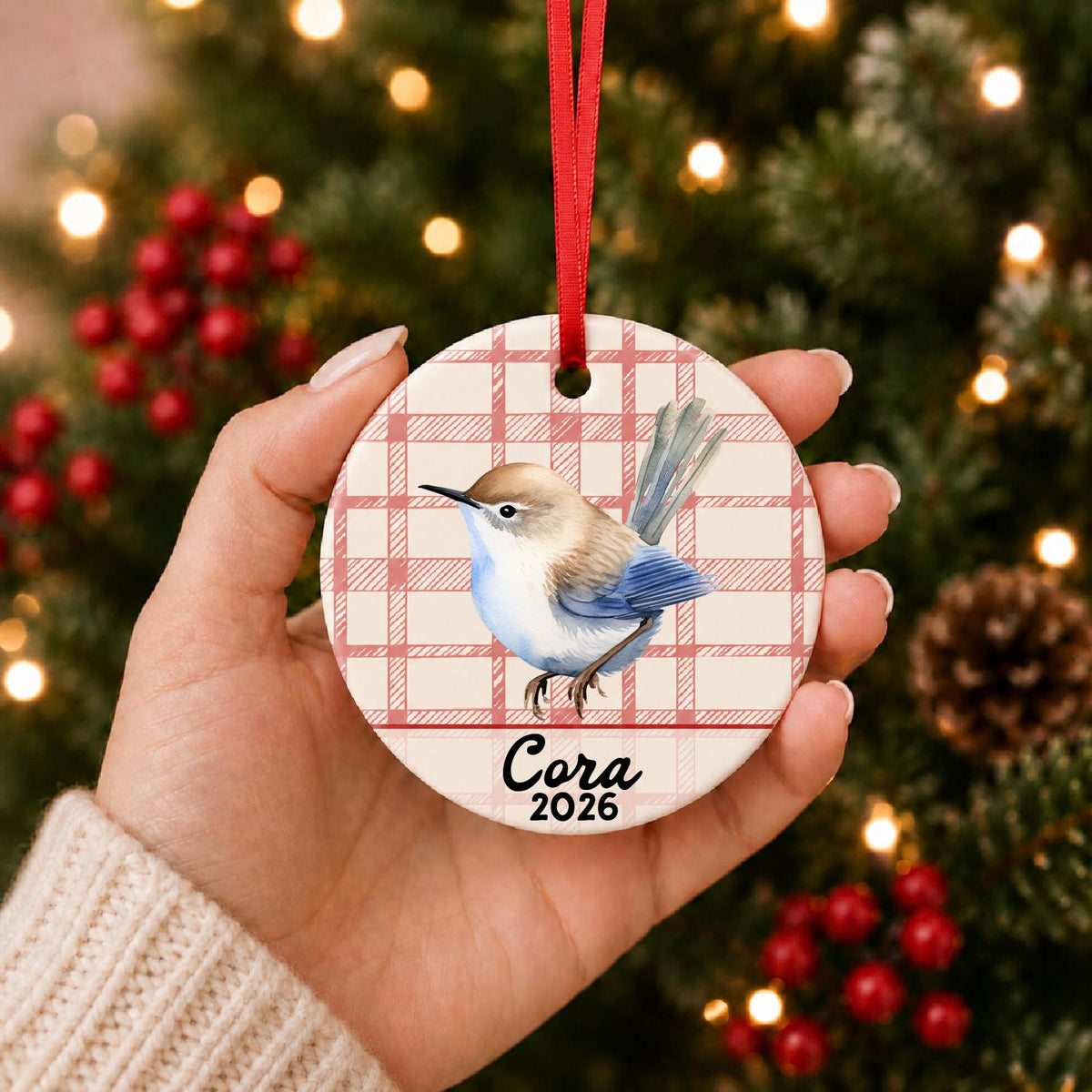 Hand holding the personalized wren ceramic ornament with name and year on red plaid in front of a Christmas tree