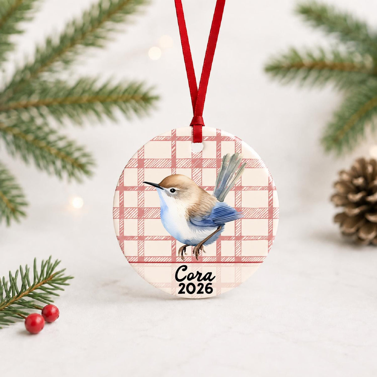 Personalized wren ceramic ornament with name and year on red plaid with red ribbon on a white surface with pine branches and pinecones