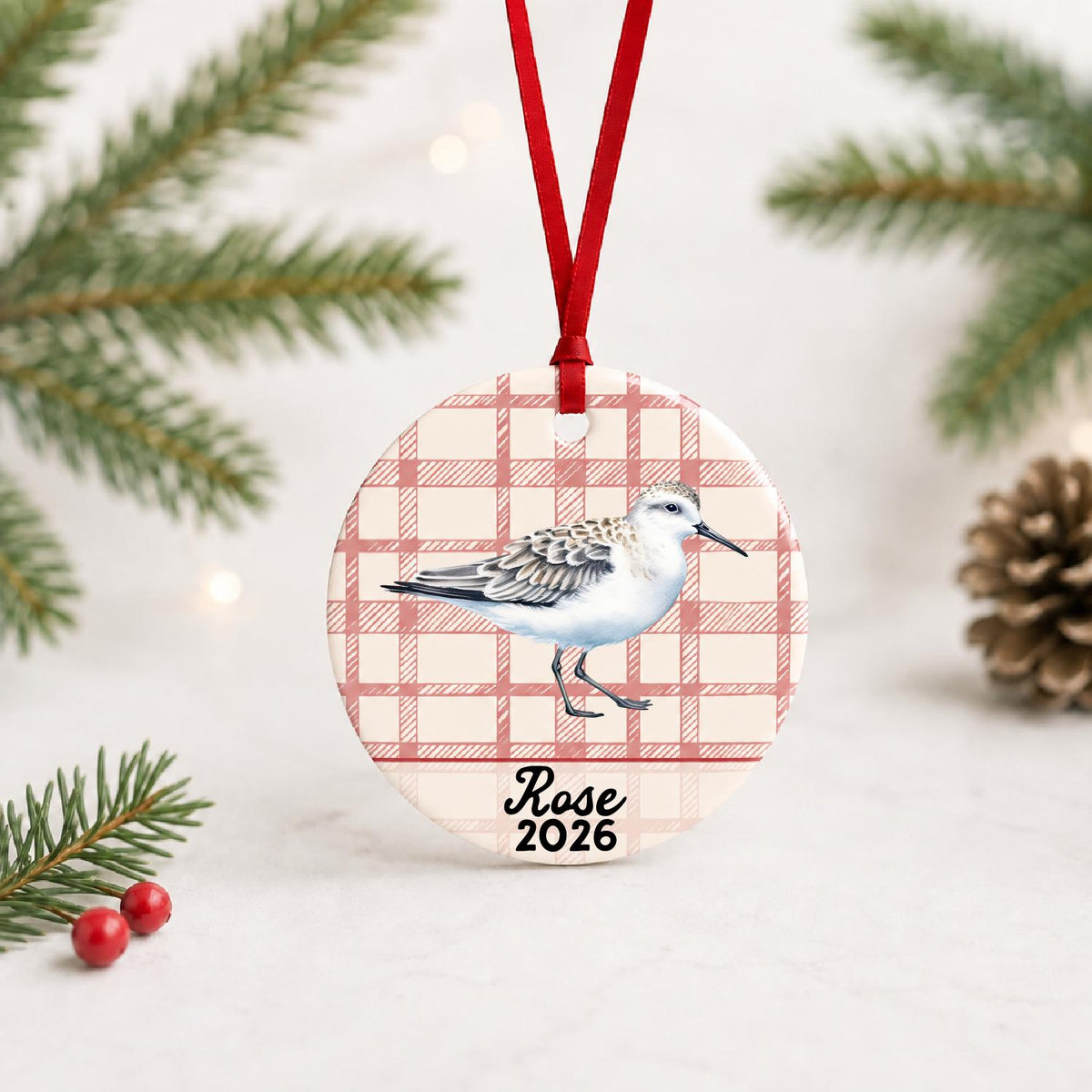 Personalized sanderling ceramic ornament with name and year on red plaid with red ribbon on a white surface with pine branches and pinecones