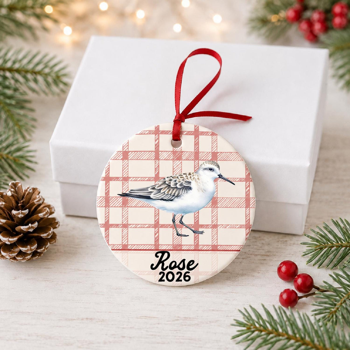 Personalized sanderling ceramic ornament with name and year on red plaid in front of a white gift box with pinecones and red berries