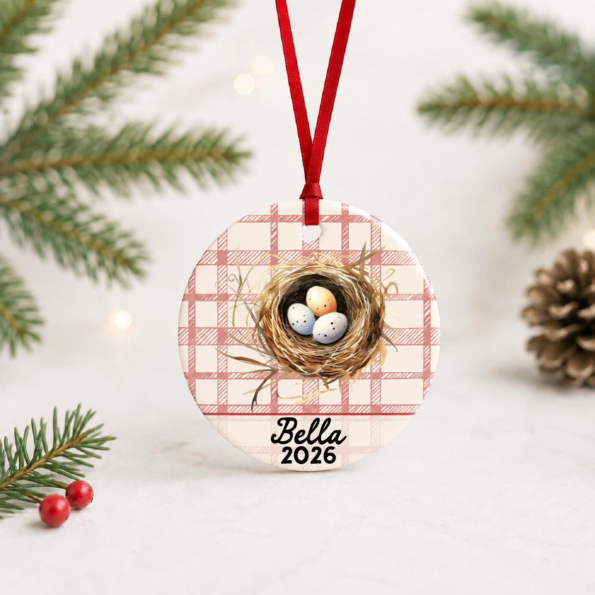 Personalized bird nest ceramic ornament with name and year on red plaid with red ribbon on a white surface with pine branches and pinecones
