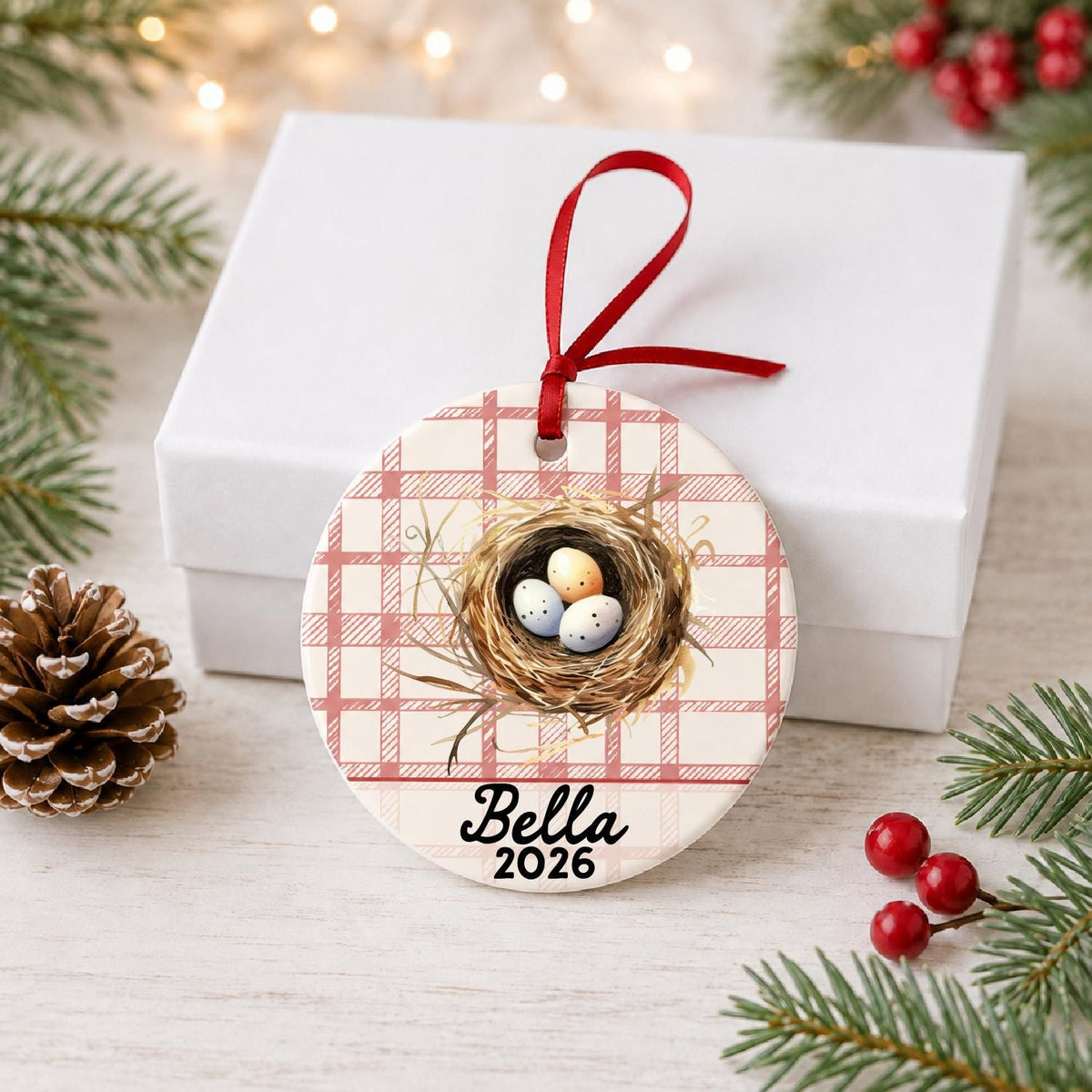 Personalized bird nest ceramic ornament with name and year on red plaid in front of a white gift box with pinecones and red berries