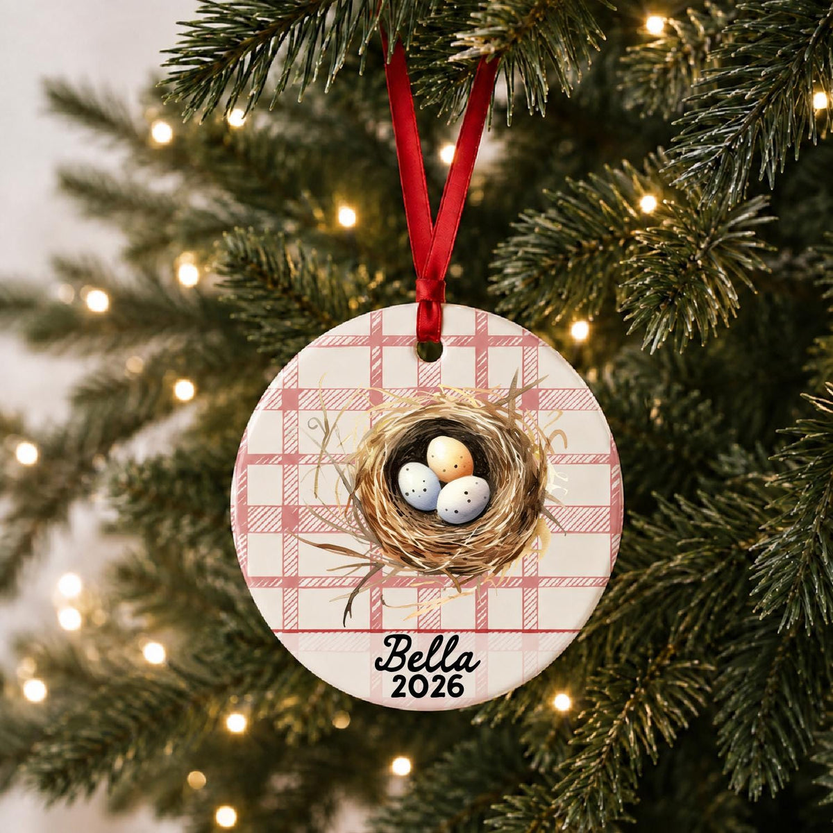 Personalized bird nest ceramic ornament with name and year on red plaid hanging on a lit Christmas tree