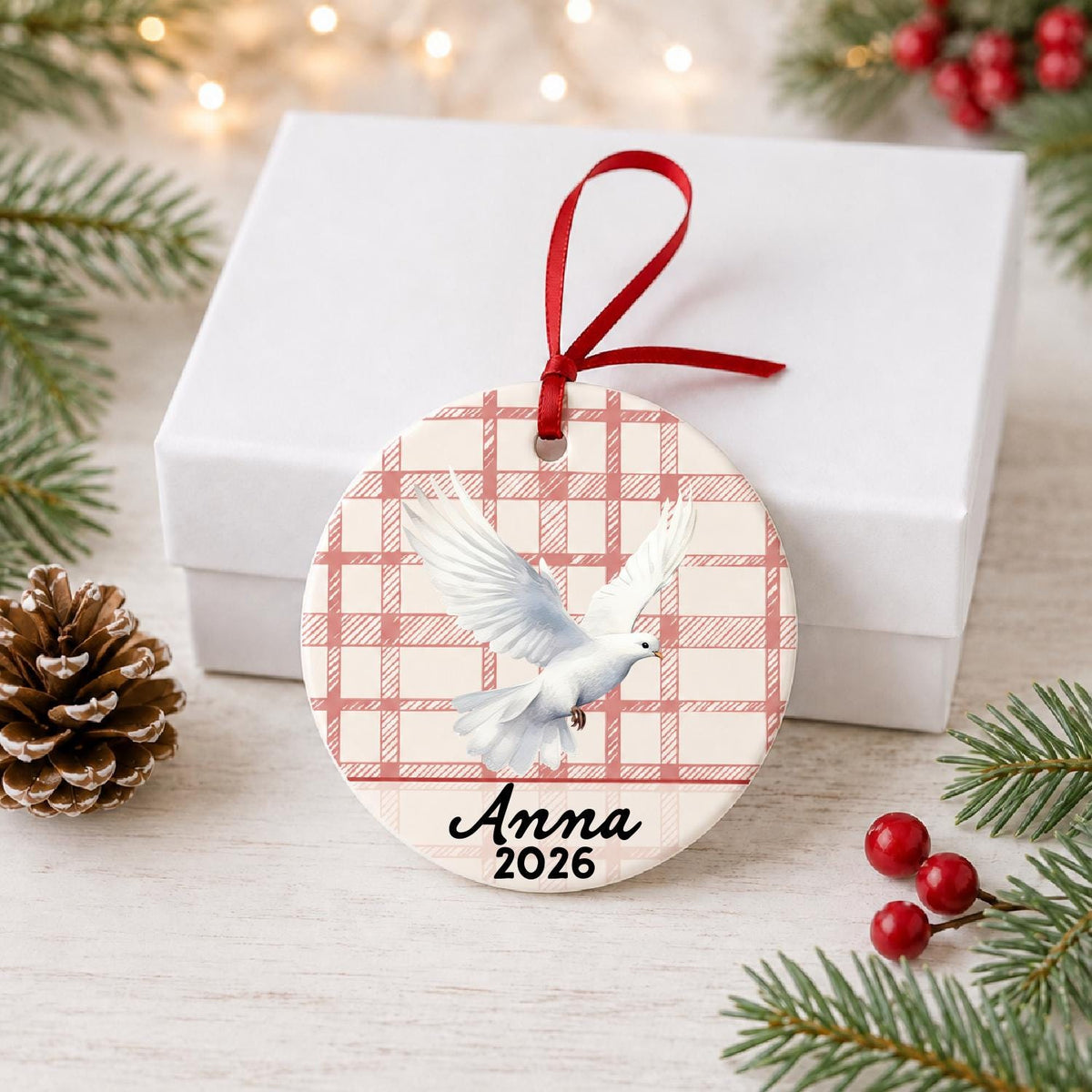 Personalized white dove ceramic ornament with name and year on red plaid in front of a white gift box with pinecones and red berries