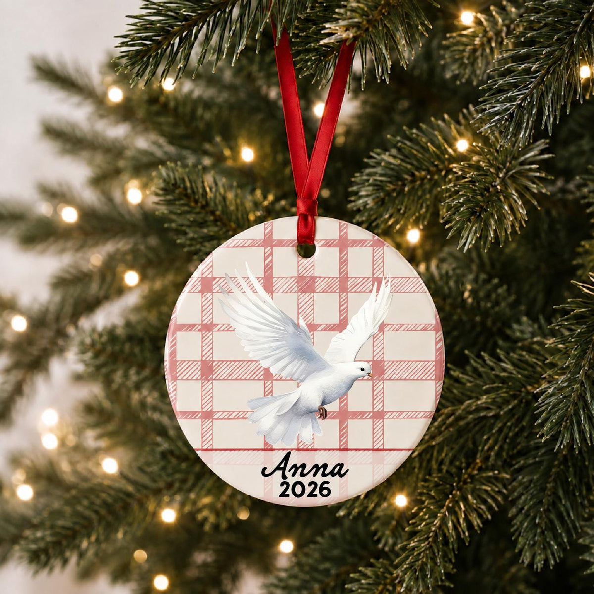 Personalized white dove ceramic ornament with name and year on red plaid hanging on a lit Christmas tree