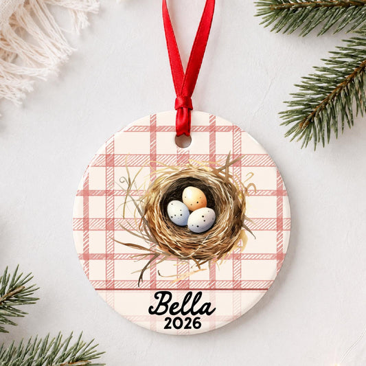 Personalized bird nest ceramic ornament with name and year on red plaid styled with pine branches on a soft white background