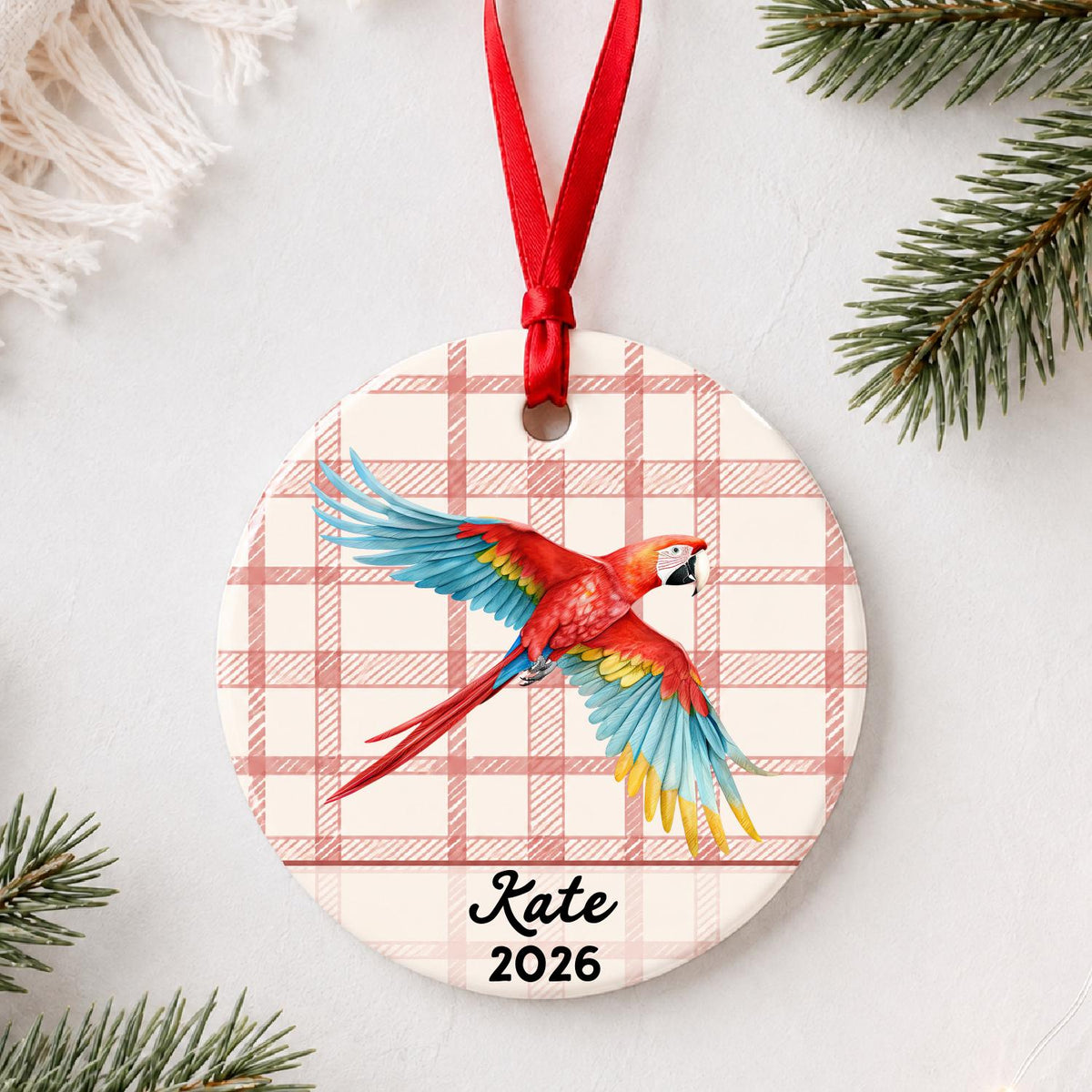 Personalized scarlet macaw ceramic ornament with name and year on red plaid styled with pine branches on a soft white background