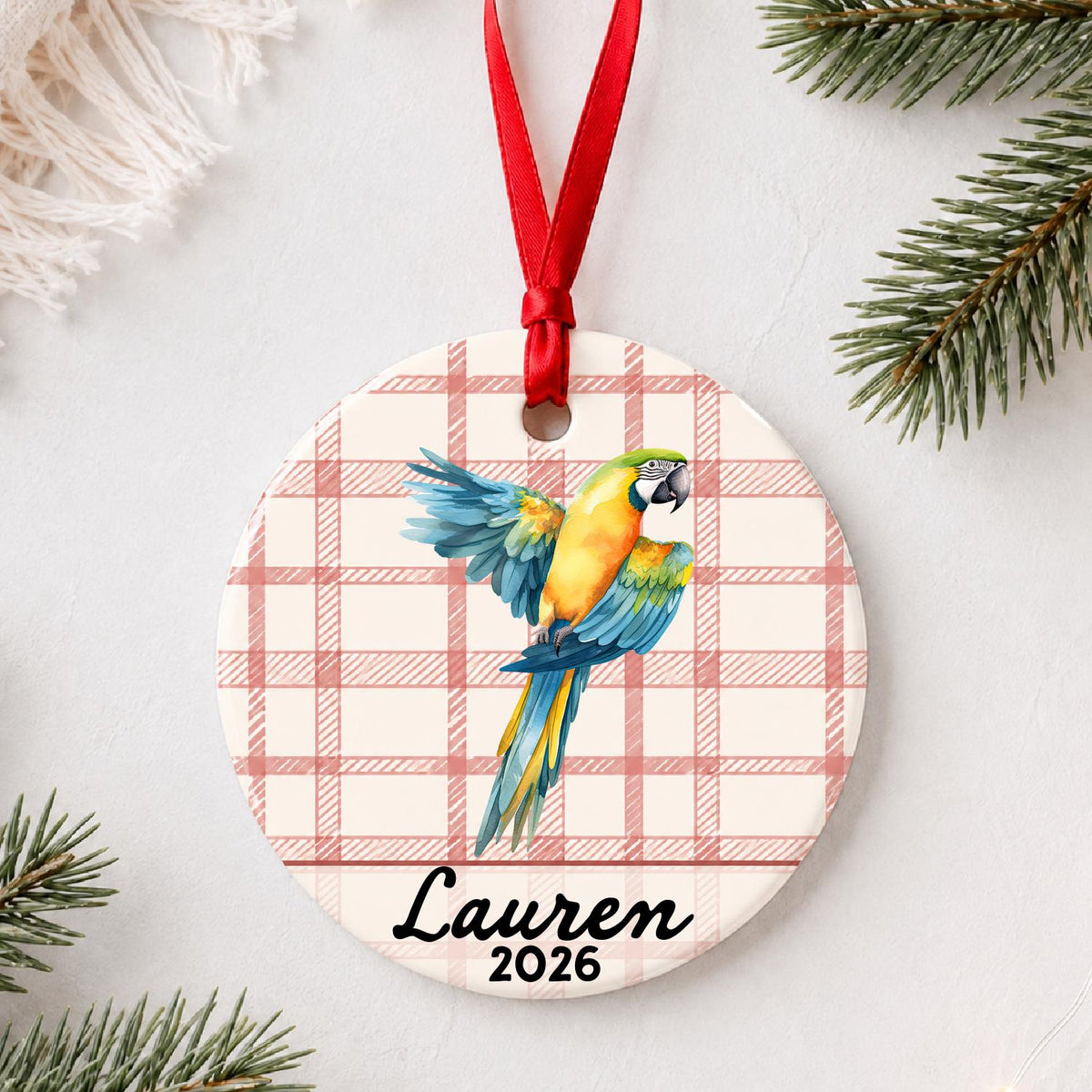 Personalized blue and gold macaw ceramic ornament with name and year on red plaid styled with pine branches on a soft white background