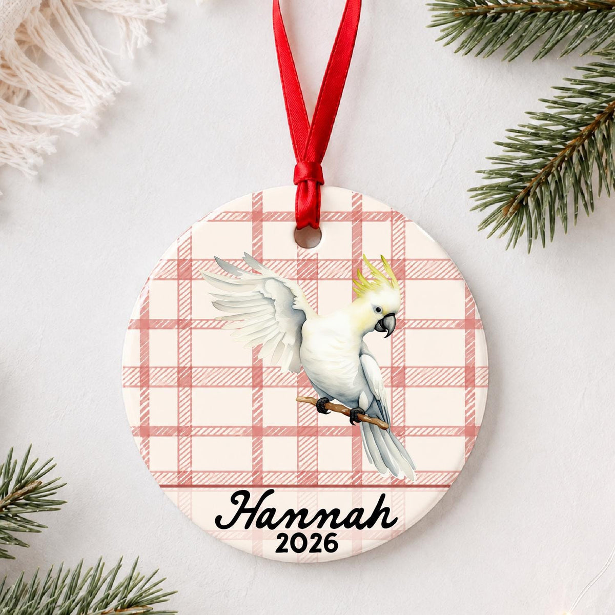 Personalized cockatoo ceramic ornament with name and year on red plaid styled with pine branches on a soft white background