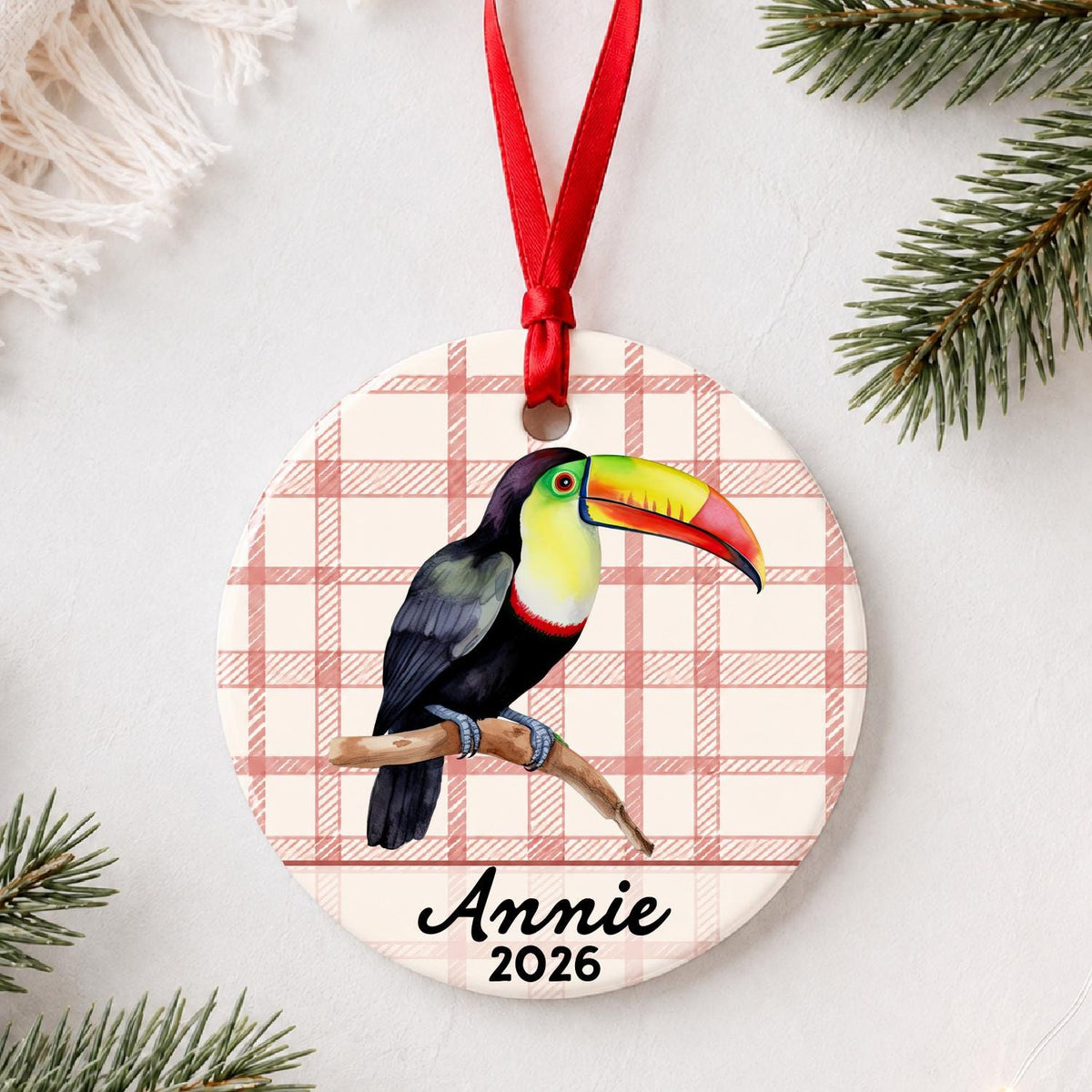 Personalized toucan ceramic ornament with name and year on red plaid styled with pine branches on a soft white background