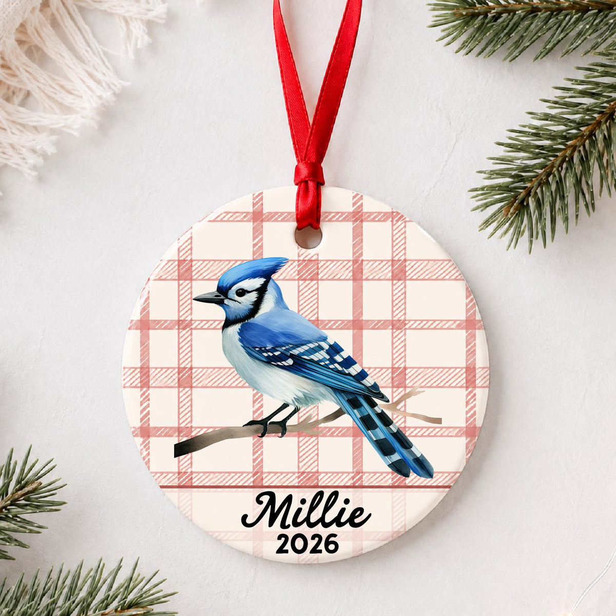 Personalized blue jay ceramic ornament with name and year on red plaid styled with pine branches on a soft white background