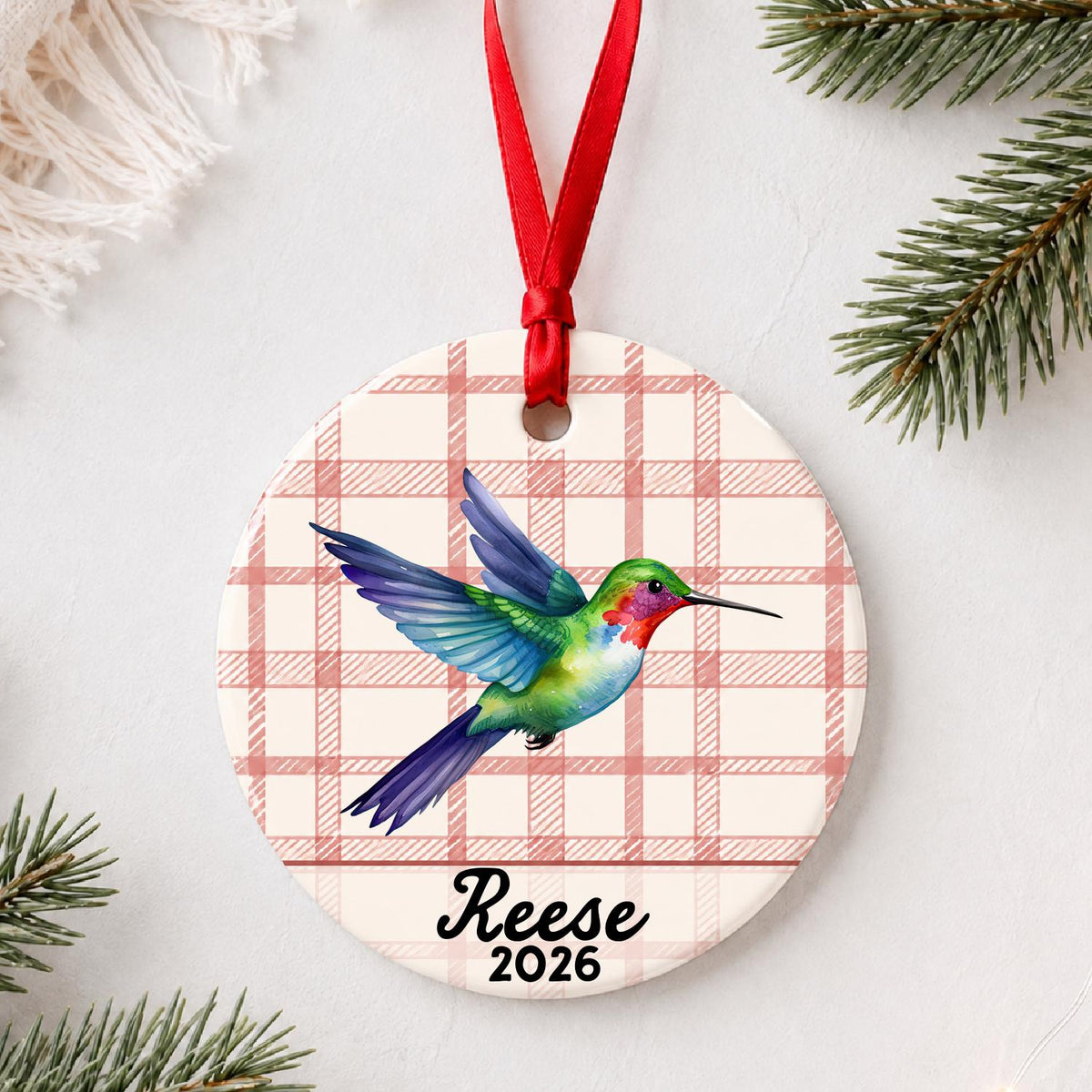 Personalized hummingbird ceramic ornament with name and year on red plaid styled with pine branches on a soft white background