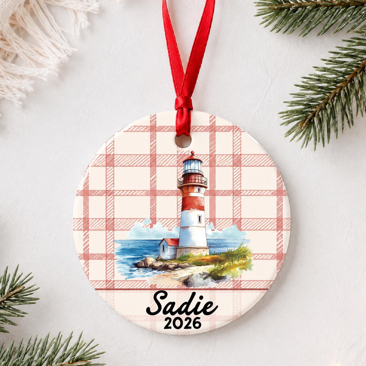Personalized beach lighthouse ceramic ornament with name and year on red plaid styled with pine branches on a soft white background