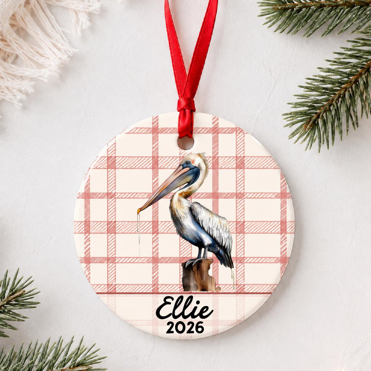 Personalized pelican on piling ceramic ornament with name and year on red plaid styled with pine branches on a soft white background