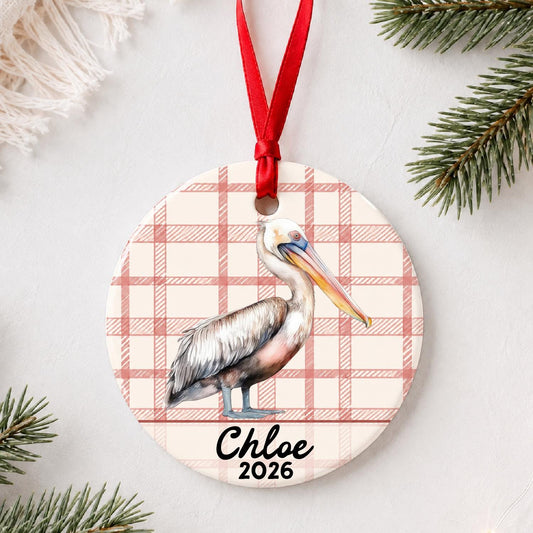 Personalized pelican ceramic ornament with name and year on red plaid styled with pine branches on a soft white background